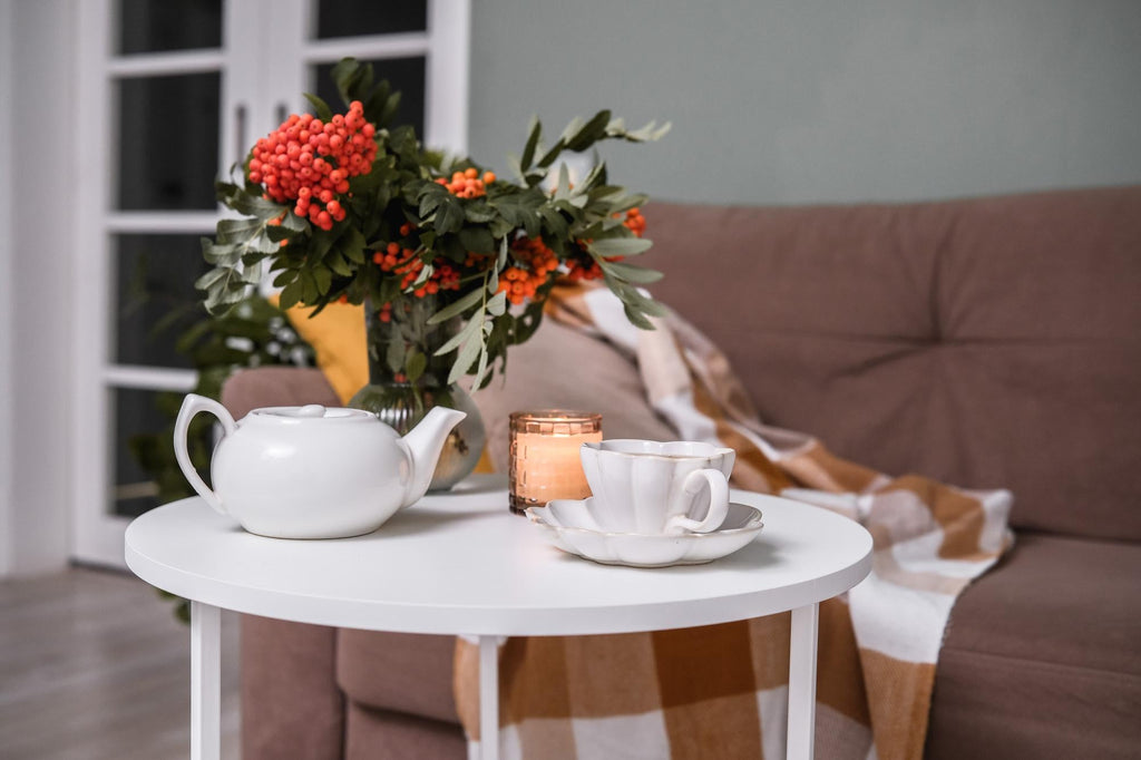White round coffee table styled with a ceramic teapot, tea cup, lit candle, and berry bouquet in a cosy living room with a brown sofa
