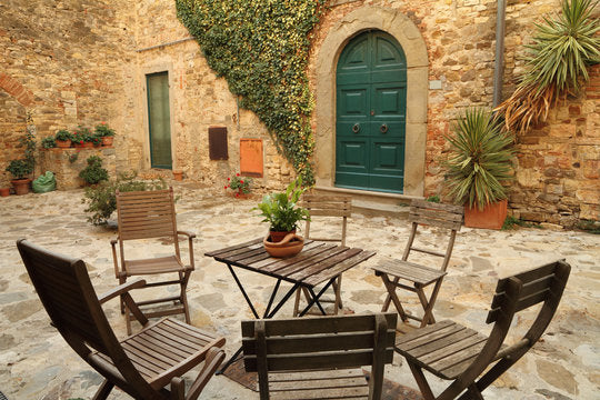 Rustic Tuscan courtyard with wooden bistro chairs and table on cobblestone patio surrounded by stone walls and climbing ivy.