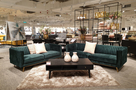 Showroom display of two teal velvet channel-back sofas with gold legs and decorative cushions, styled around a dark wood coffee table on a shag rug.