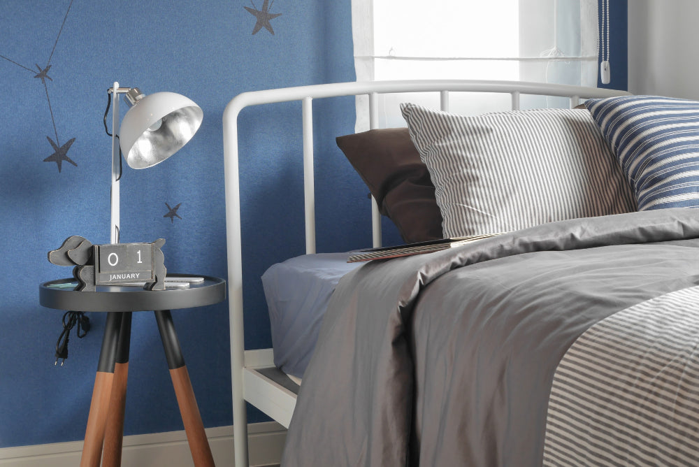 Blue teen bedroom with white metal bed frame, grey striped bedding, and industrial desk lamp on black tripod side table.