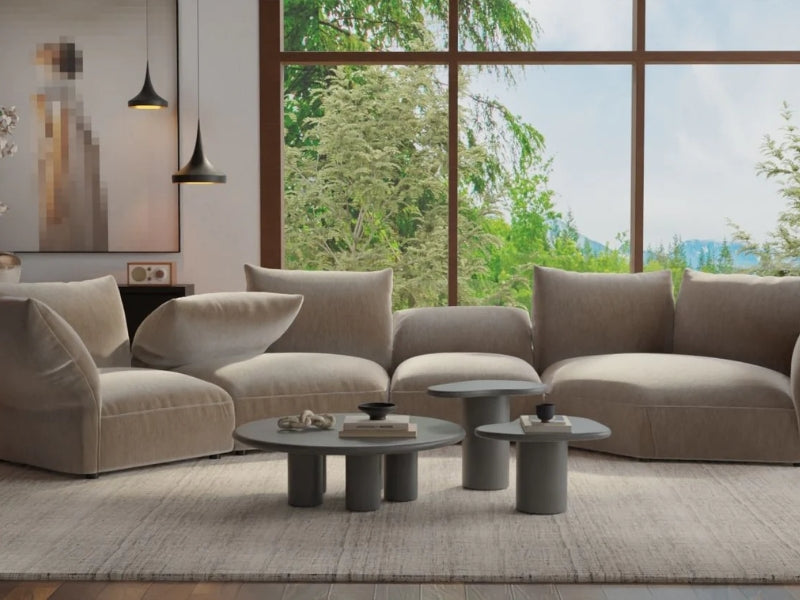 Curved beige sectional sofa with plush cushions in a modern living room, paired with round nesting coffee tables, pendant lighting, and large windows overlooking greenery.