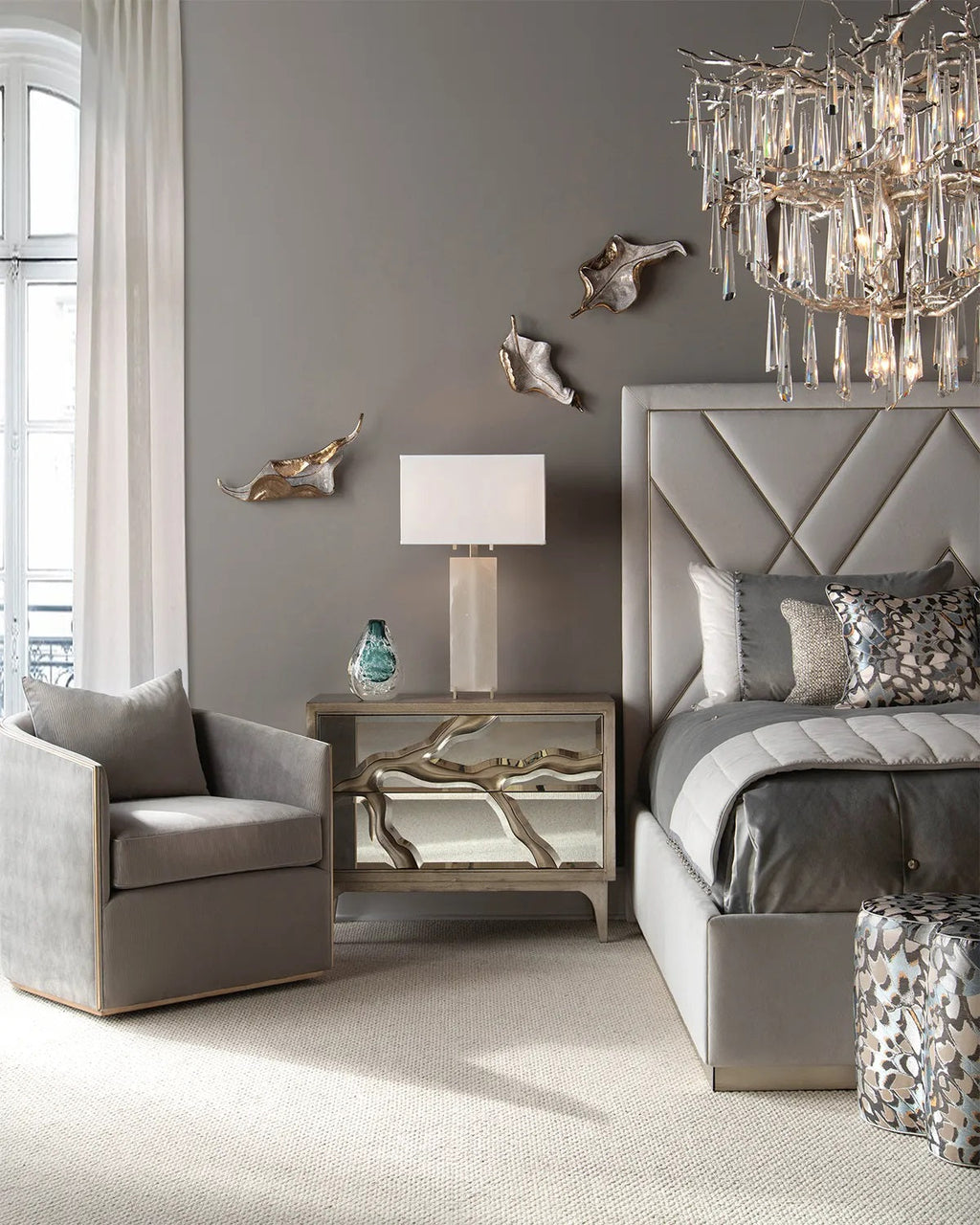 Grey upholstered bed with diamond-pattern gold trim headboard beside mirrored nightstand with organic brass hardware in glam bedroom with crystal chandelier