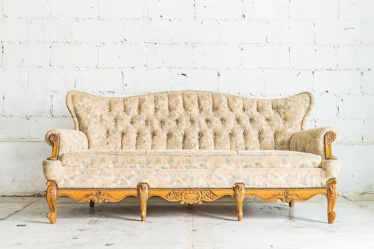 Vintage French provincial sofa with tufted floral upholstery and carved oak wood frame against white brick wall.