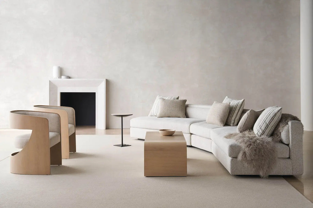 Curved beige sectional sofa with textured pillows and sheepskin throw in a minimalist living room with a stone fireplace, wooden barrel chairs, and a cube coffee table