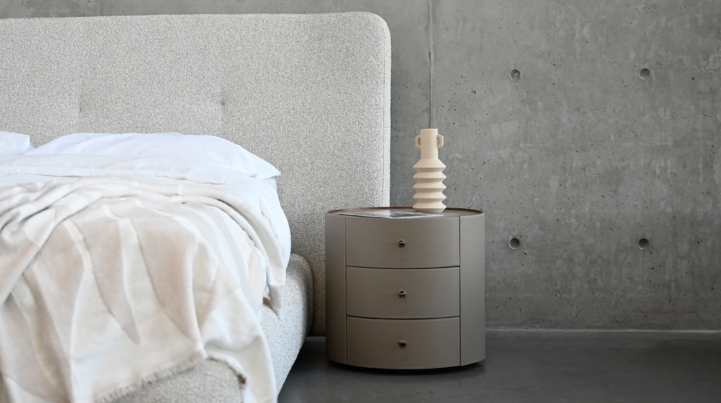 Cylindrical three-drawer leather nightstand in taupe grey with rose gold knobs beside grey upholstered bed against concrete wall