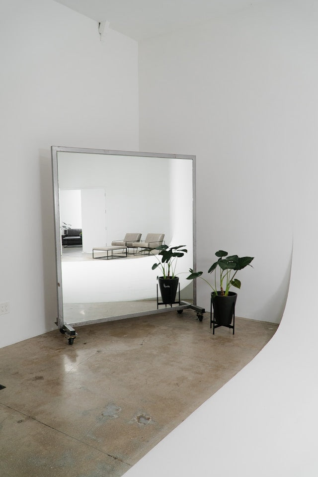 Large freestanding mirror on wheels in minimalist white photography studio with potted tropical plants on concrete floor.