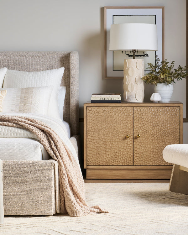 A styled bedroom scene featuring a textured light wood nightstand with two doors and small gold handles, styled with a white lamp, books, and a small plant. The nightstand is positioned next to a bed with neutral beige and white bedding.