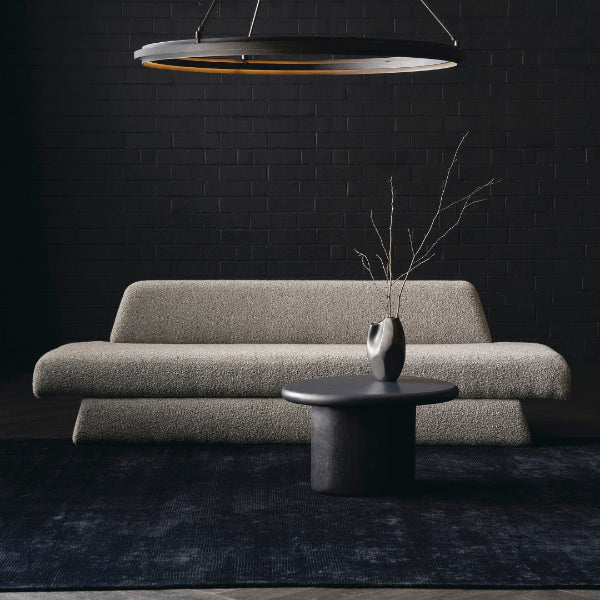 A modern beige bouclé sofa in a dark, stylish living room setting with a black brick wall, a dark round coffee table, a dark blue rug, and a hanging ring light.