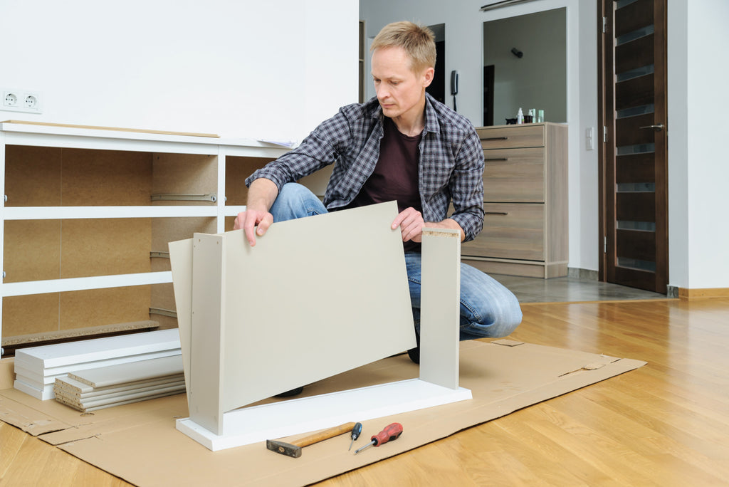 Man assembling a flat-pack TV stand on a hardwood floor with screwdrivers and a hammer nearby, in a bright modern living room.