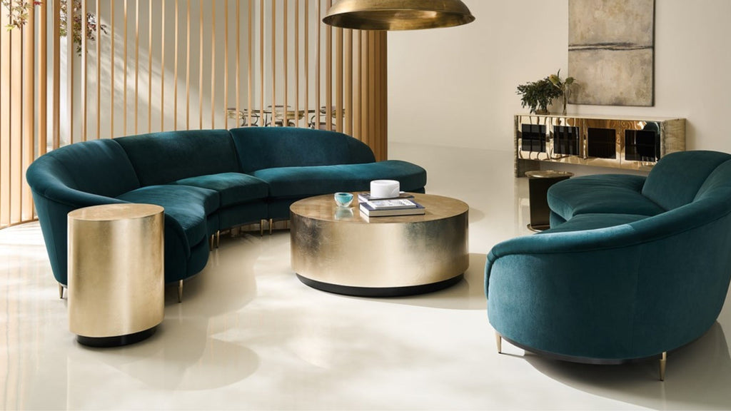 A modern living room setup featuring a curved teal velvet sectional sofa, a matching rounded armchair, and metallic gold furniture including a side table and round coffee table.