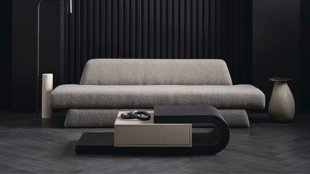A modern light gray textured sofa in a dark, minimalist living room setting with a black and wood coffee table in front.