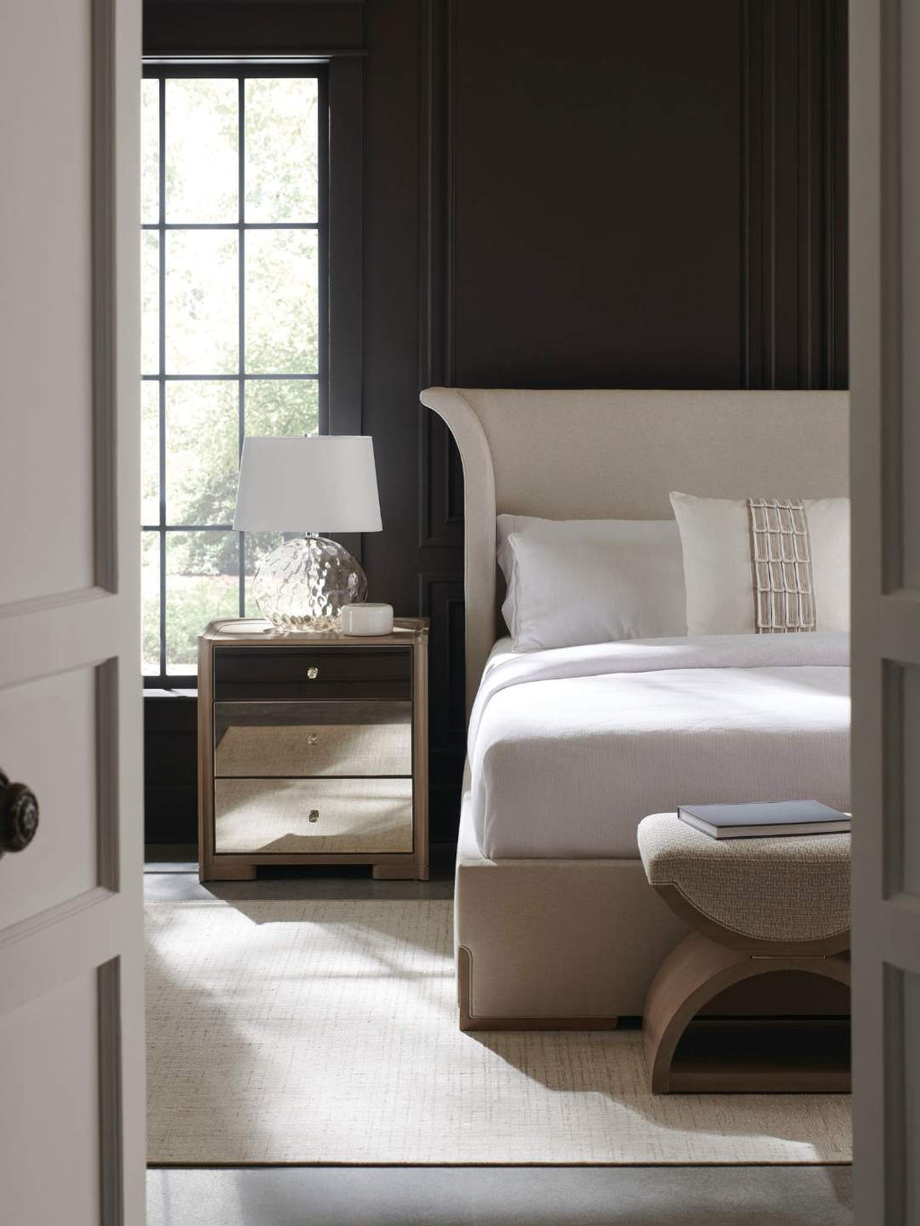 A styled bedroom interior showing a bed with white bedding, a beige upholstered headboard and footboard, a mirrored nightstand with a lamp, and a dark paneled wall. The scene is viewed through an open doorway, with natural light coming from the window.