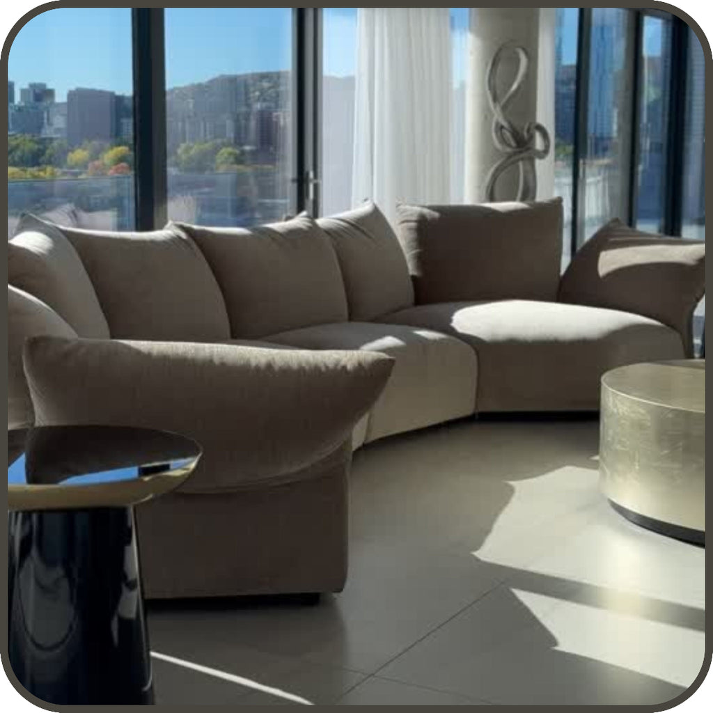 A curved modular upholstered sofa in a light taupe-beige fabric, styled in a modern living room with large floor-to-ceiling windows, a round metallic coffee table, and a dark side table.