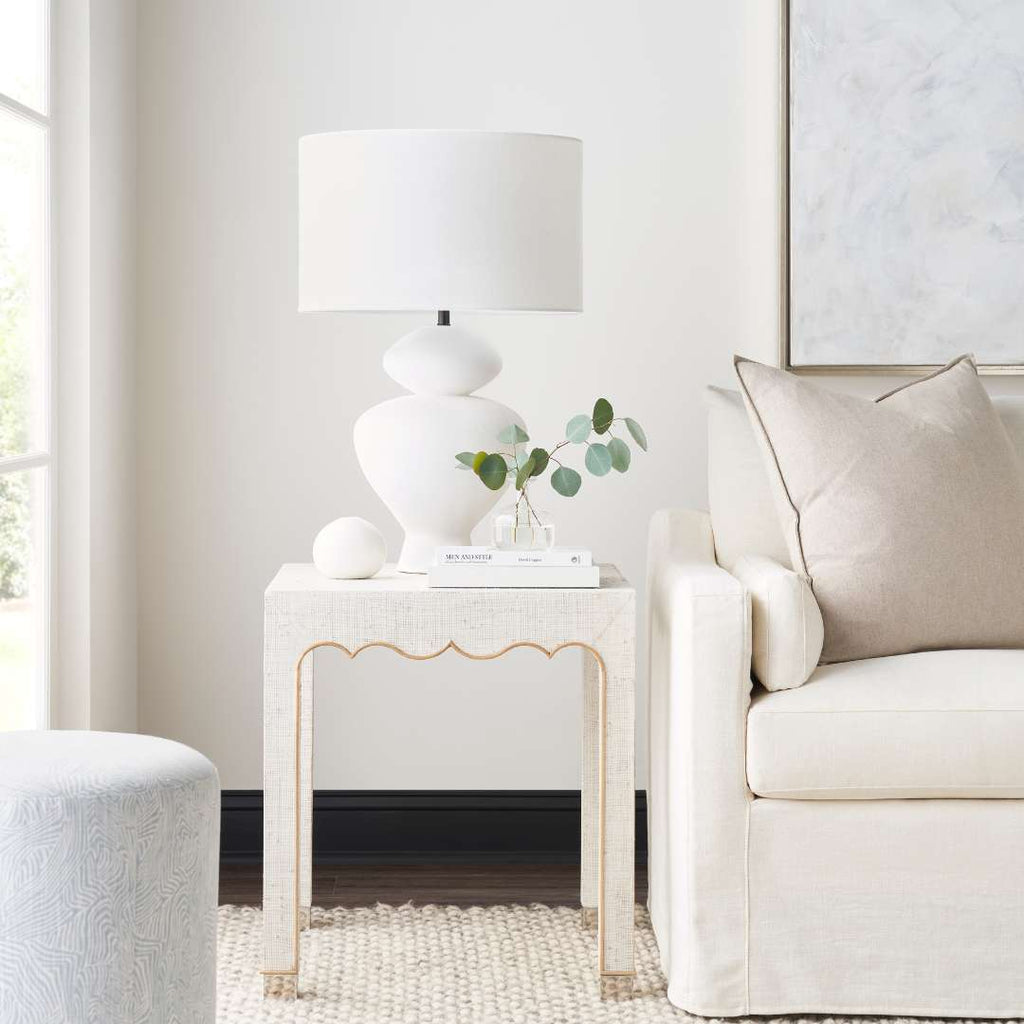 A white ceramic table lamp with a drum shade sits on a white textured side table in a bright, modern living room next to a cream-colored armchair.