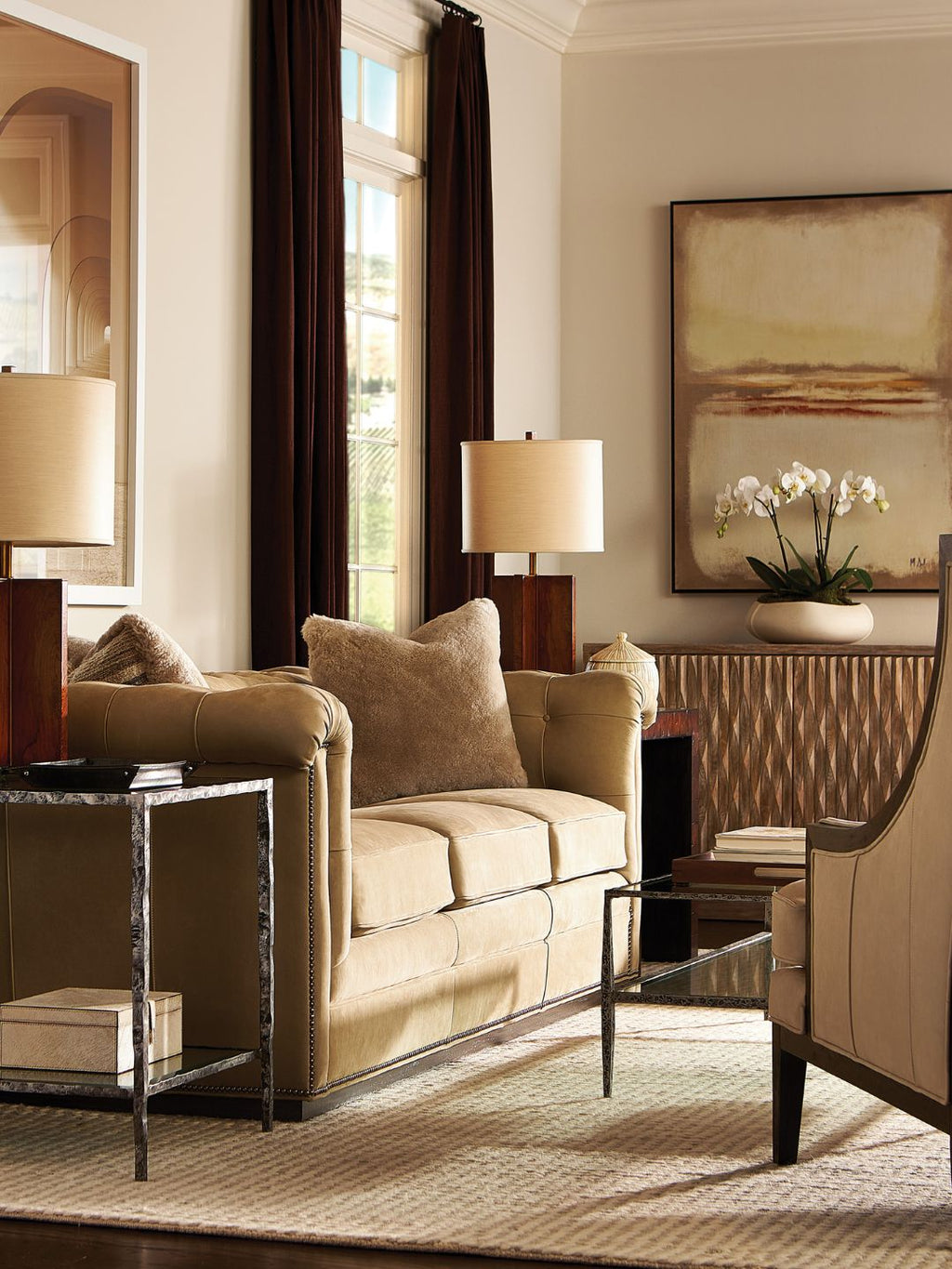 A beige upholstered loveseat in a styled living room with dark brown curtains, matching side tables, table lamps, a patterned rug, and wall art.