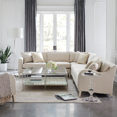 A modern living room featuring a large U-shaped sectional sofa upholstered in warm cream fabric, styled with matching throw pillows, a glass coffee table, and a clear acrylic side table in front of a large window with dark gray curtains.