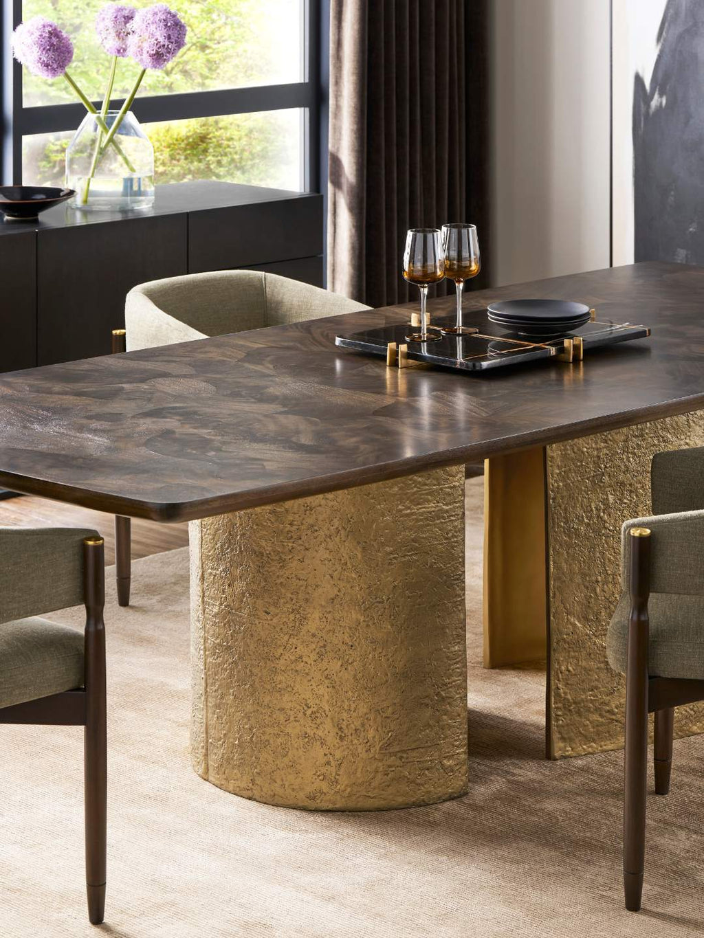 A modern dining table with a dark wood top and textured gold cylindrical pedestal base, surrounded by upholstered dining chairs in a styled interior room.