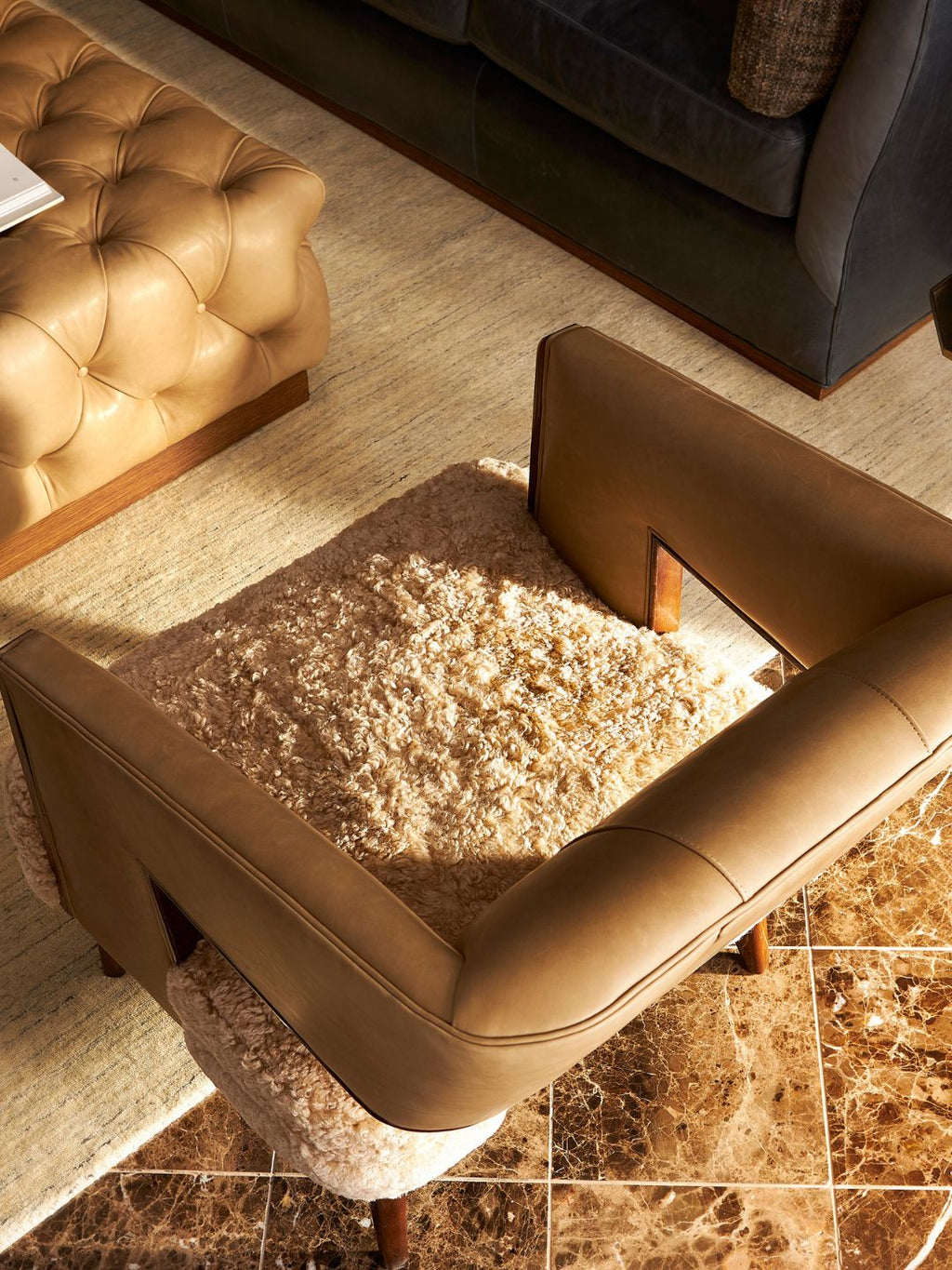 A brown leather armchair with a thick beige shag rug on its seat, styled in a modern living room setting with a tufted leather ottoman and a dark gray sofa in the background.