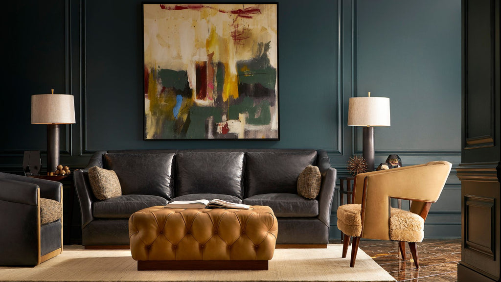 A styled living room featuring a dark charcoal leather sofa, a tufted mustard-brown leather ottoman, a beige armchair, and a large abstract painting on a dark teal paneled wall.