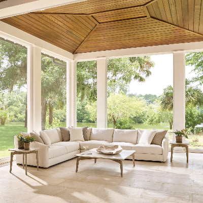 A large sectional sofa in off-white and beige tones arranged on a covered porch with a wood ceiling, light stone floor, and large windows overlooking a lush green outdoor lawn.