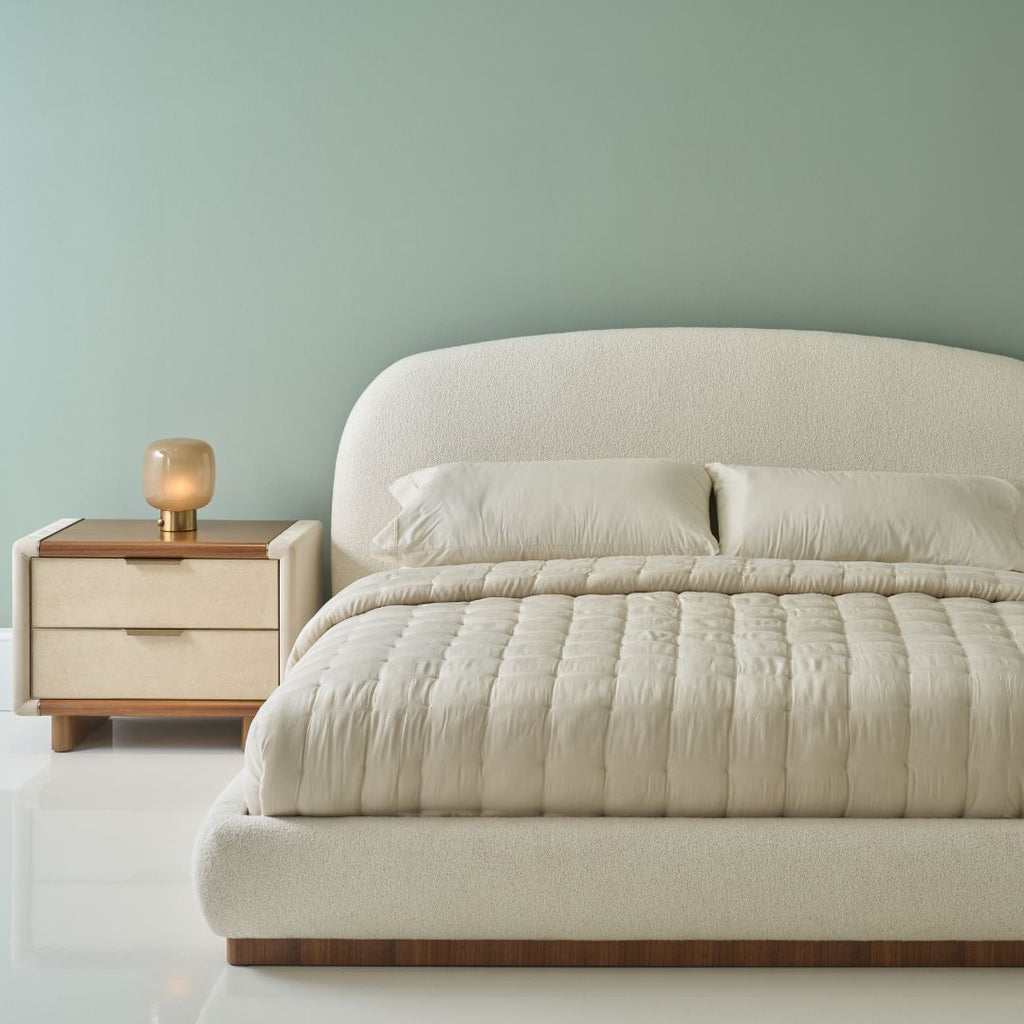 A modern upholstered bed with a rounded headboard and matching quilted cream bedding, shown beside a two-tone nightstand against a soft sage green wall.