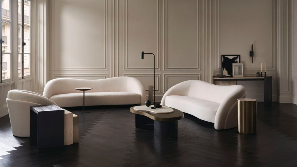 A luxury living room featuring two modern curved white sofas, a matching armchair, and sculptural dark wood coffee tables against elegant cream paneled walls and dark wood floors.
