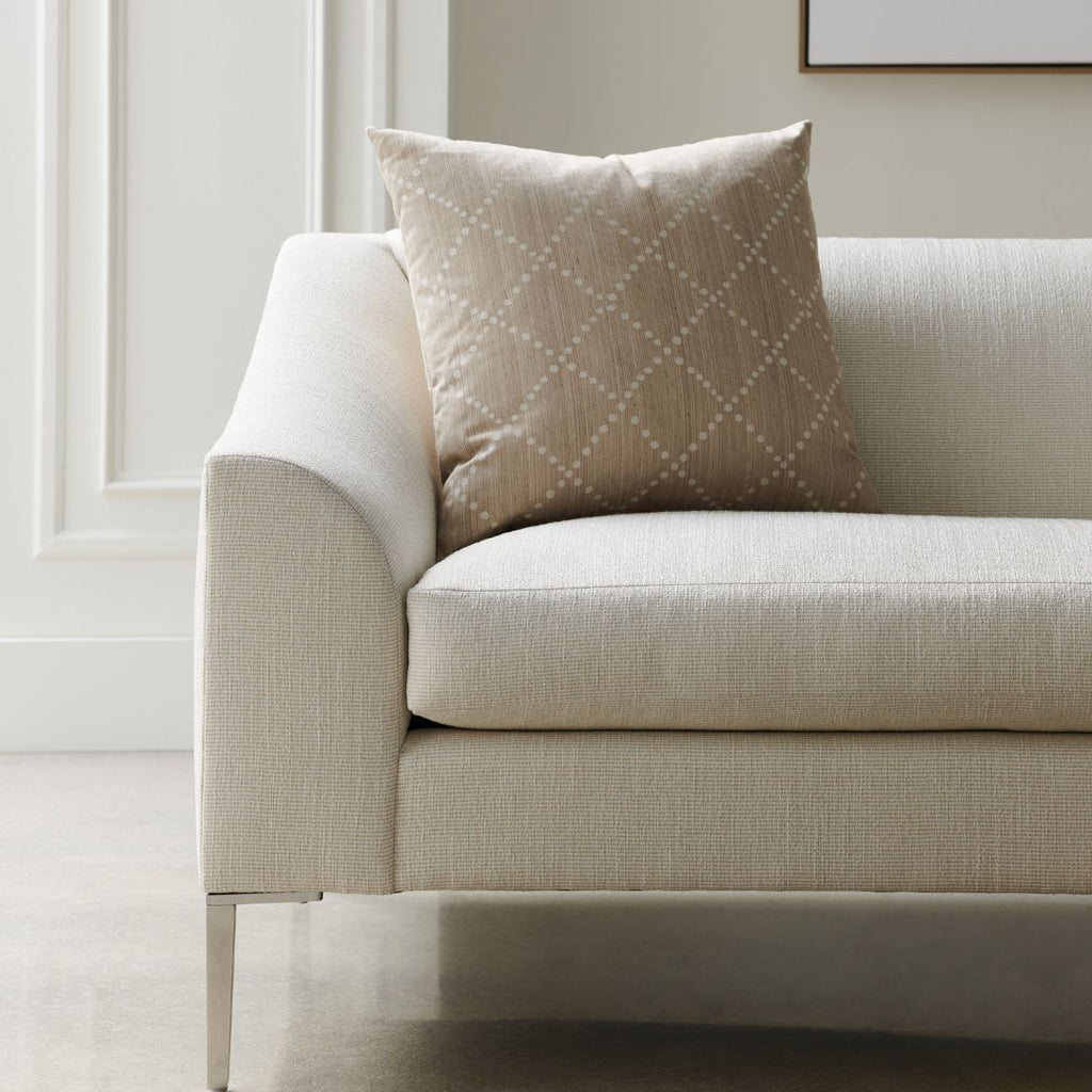 A close-up of a modern sofa in light beige textured fabric with a matching beige throw pillow featuring a subtle diamond pattern, set in a bright, minimalist room.