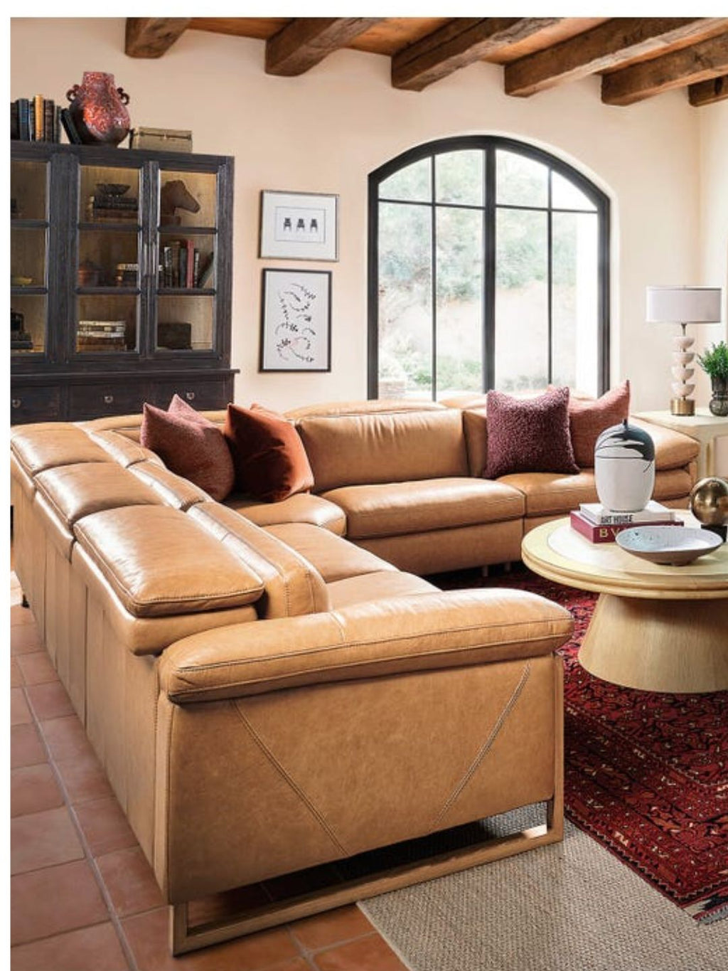 A tan leather sectional sofa with matching accent pillows in a rustic living room setting featuring a wood cabinet, arched window, and terracotta tile floor.