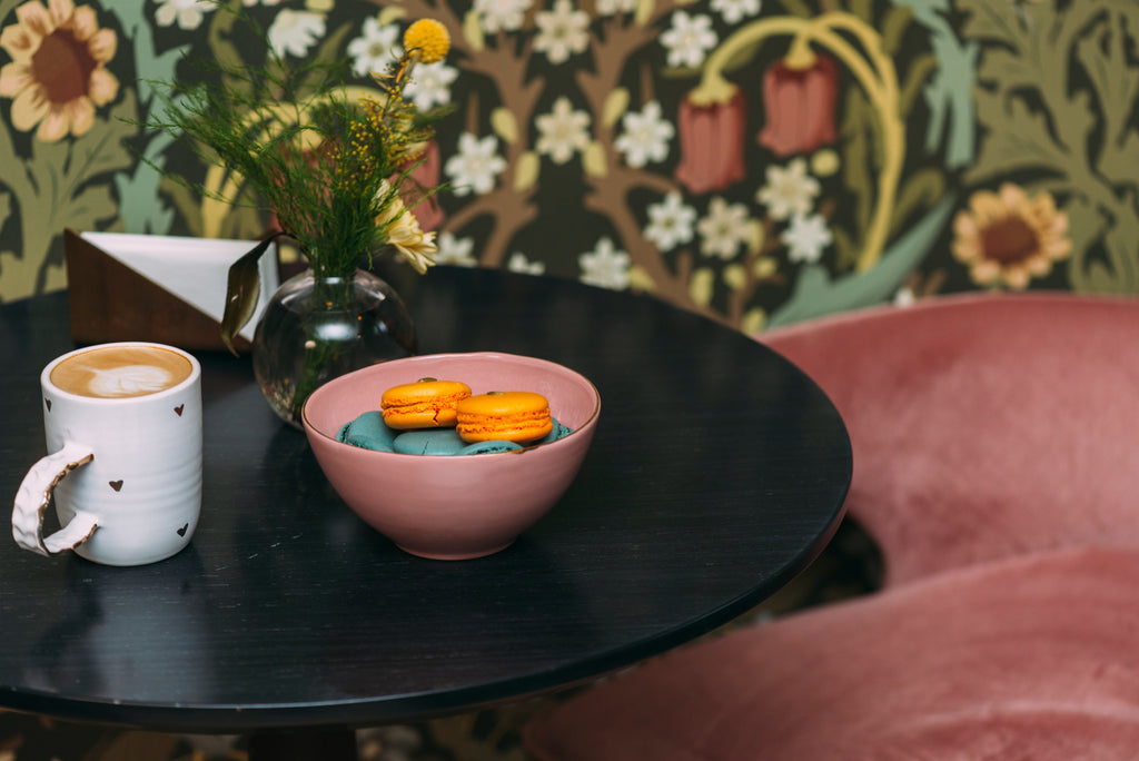 Round black side table styled with a latte, macarons in a pink bowl, and wildflowers against a bold botanical wallpaper.