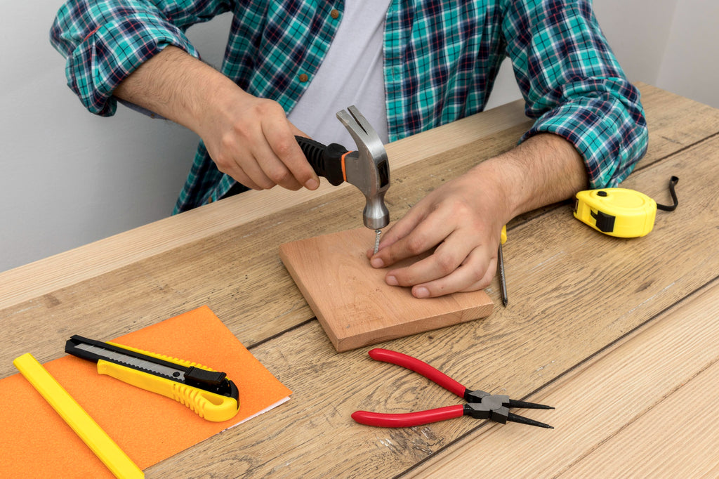 Person hammering a nail into a wooden board on a workbench, surrounded by a tape measure, pliers, and utility knife.