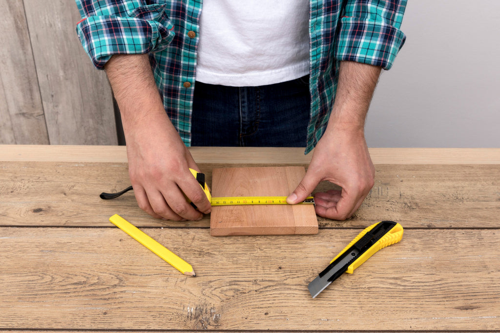 Person using a yellow tape measure to size a wooden board on a workbench, with a pencil and utility knife alongside.