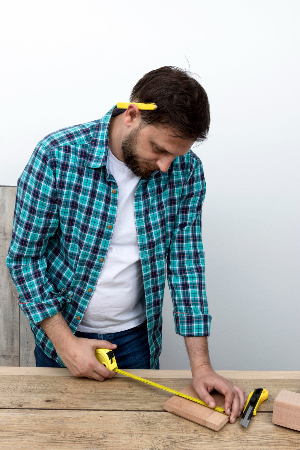 Man measuring a wooden board with a tape measure while sizing furniture, pencil tucked behind ear, against a neutral background.