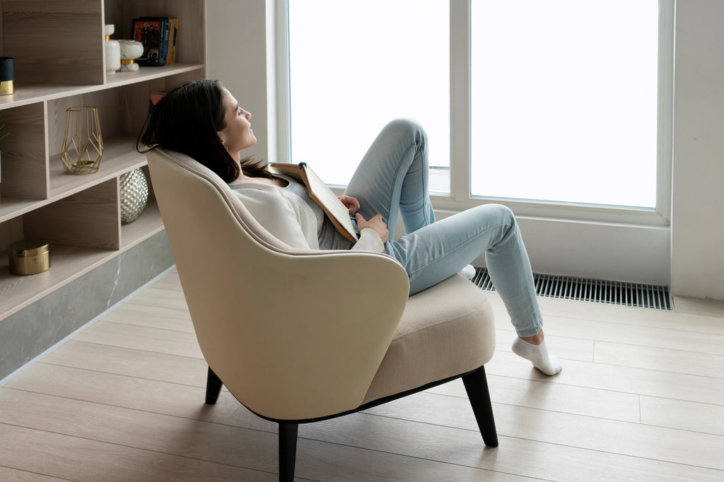 Woman relaxing in a cream upholstered accent chair, legs draped over the armrest, book in hand, beside a bright living room window.