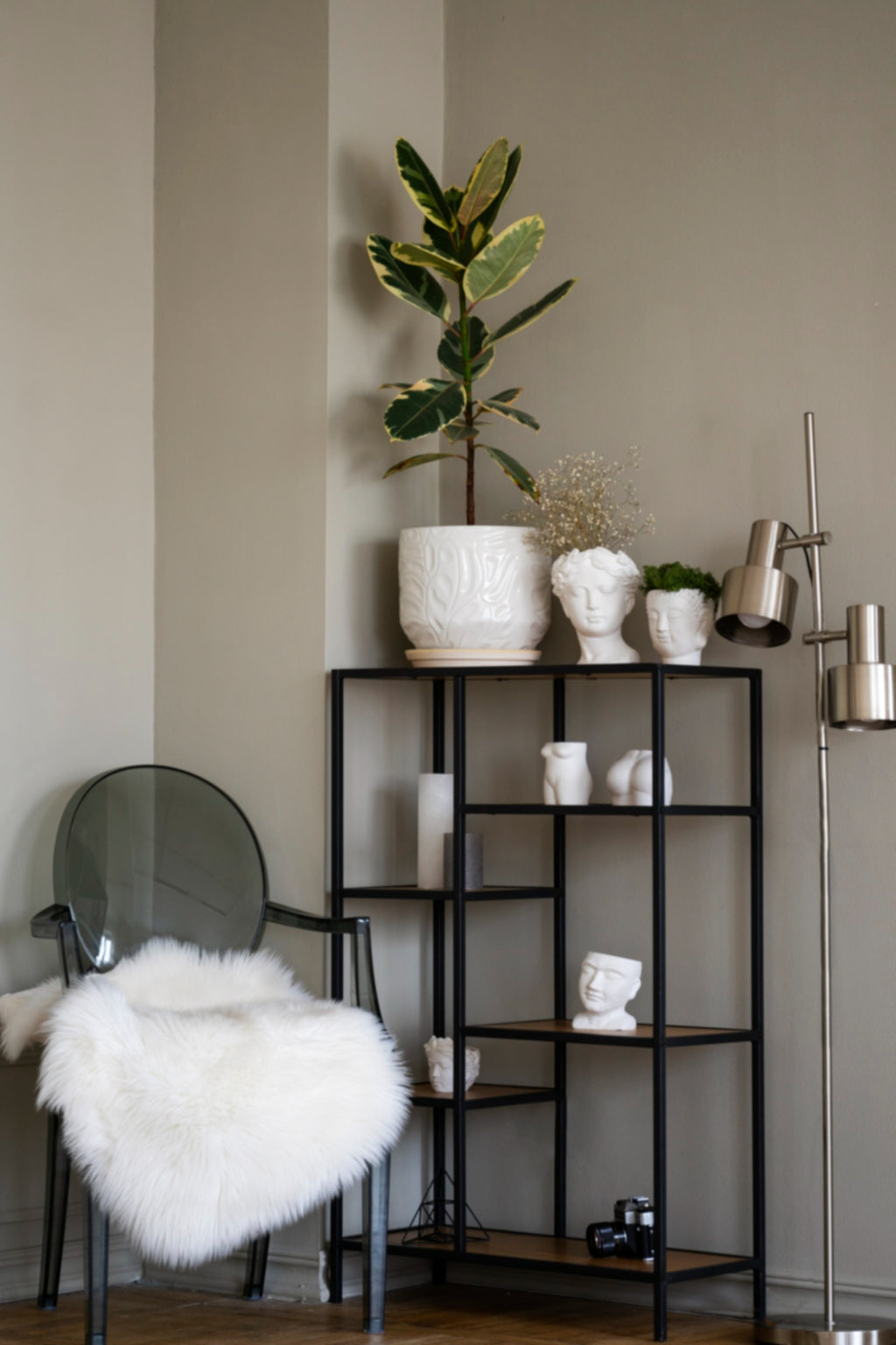 Transparent acrylic accent chair with faux fur throw beside a black metal shelving unit styled with ceramic planters and a fiddle leaf fig.