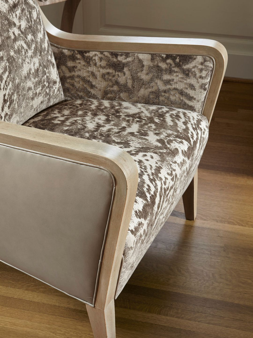 A close-up of an armchair with a beige and brown mottled patterned velvet upholstery and a light natural wood frame, set on a polished hardwood floor.