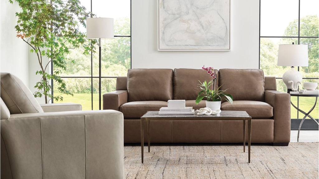A brown three-seat sofa in a modern living room setting, styled with a matching coffee table, a beige armchair, and large windows in the background.