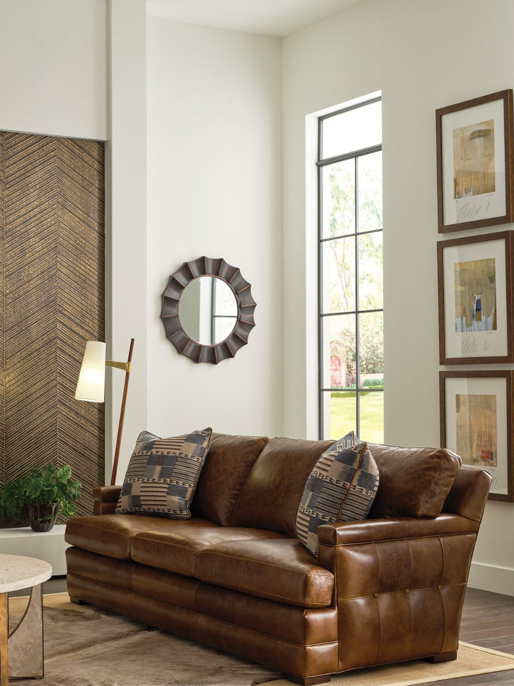 A brown leather loveseat sofa in a modern living room setting, styled with two patterned throw pillows, a floor lamp, a decorative mirror, and framed wall art.