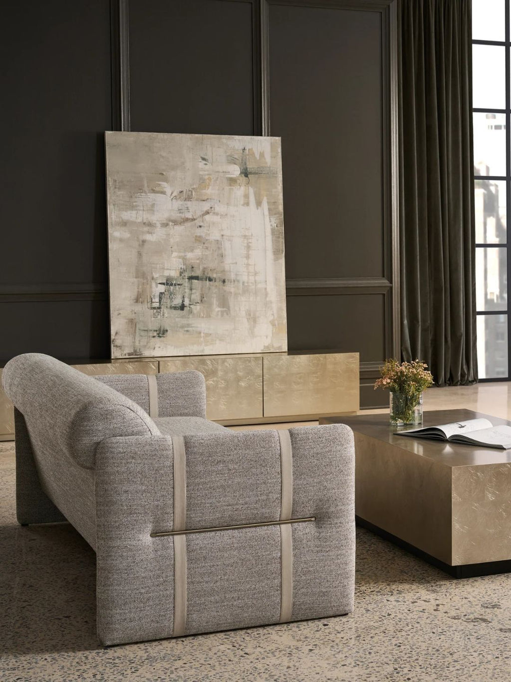 A modern light gray upholstered chaise lounge chair in a styled living room setting with a marble coffee table, dark paneled wall, and abstract wall art.