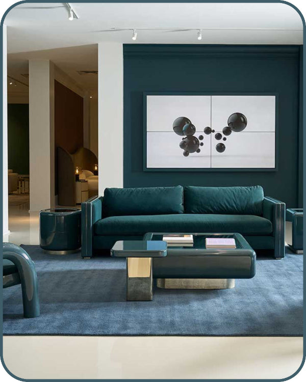 A modern living room setup featuring a dark teal velvet sofa, a matching dark teal coffee table with a glass top, and a blue area rug. The sofa is set against a dark teal accent wall with a large abstract black and white wall art piece above it.
