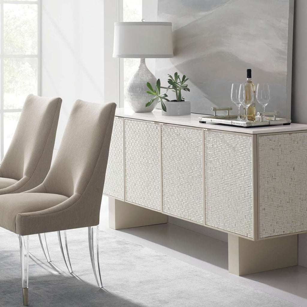 A modern sideboard or credenza in a bright dining room, styled with a textured white lamp, small potted plants, and wine glasses. Two upholstered dining chairs are positioned in the foreground on a light gray rug.