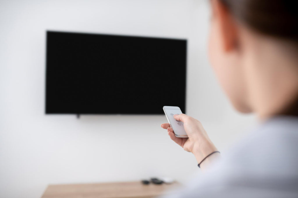 Person pointing a remote at a wall-mounted flat-screen TV in a minimalist white living room, viewed from behind.