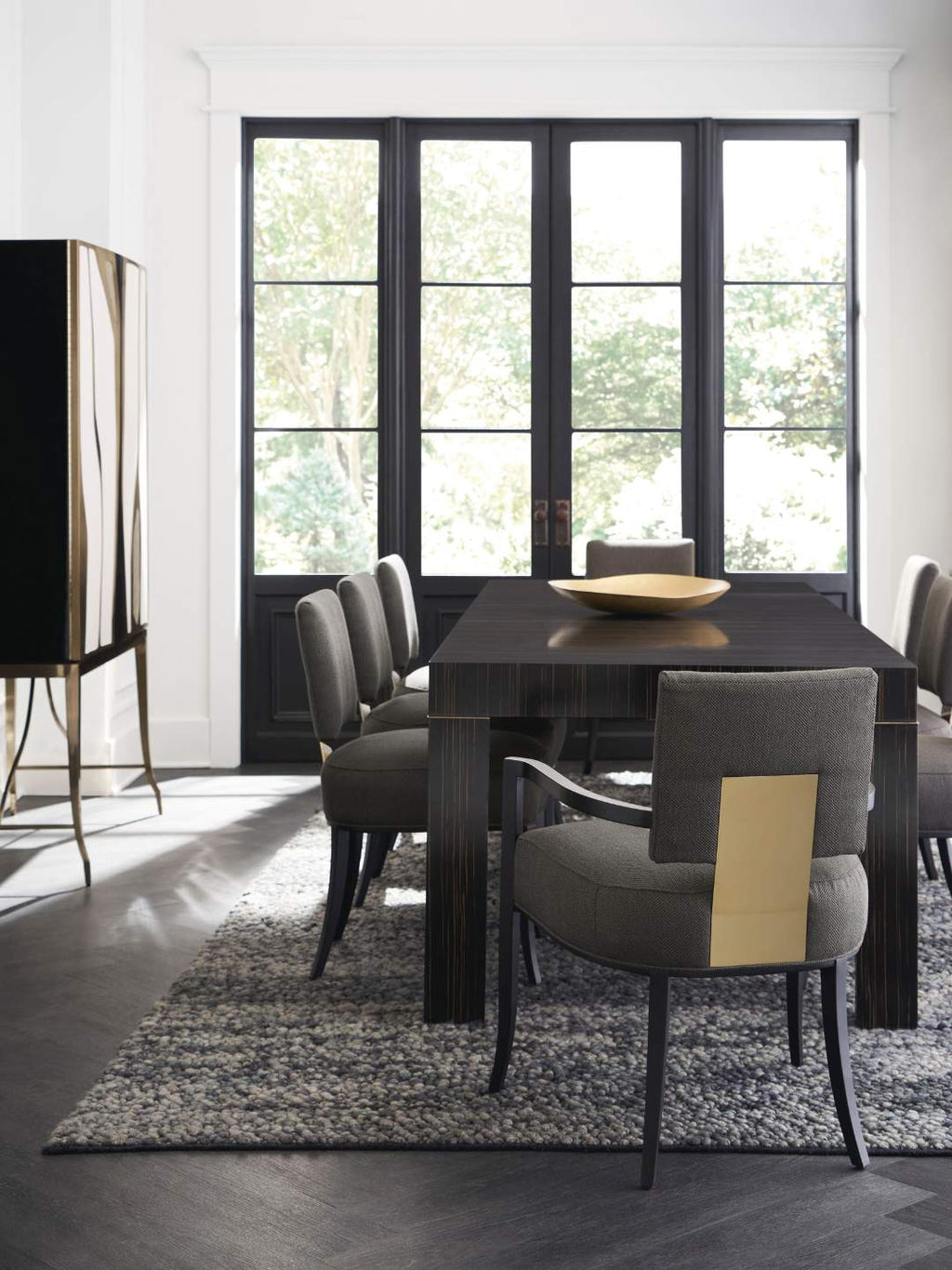 A modern dining room setup featuring a dark wood rectangular table surrounded by gray upholstered dining chairs with gold accent panels on the backrests, set on a textured gray and white patterned rug in front of black-framed glass doors.