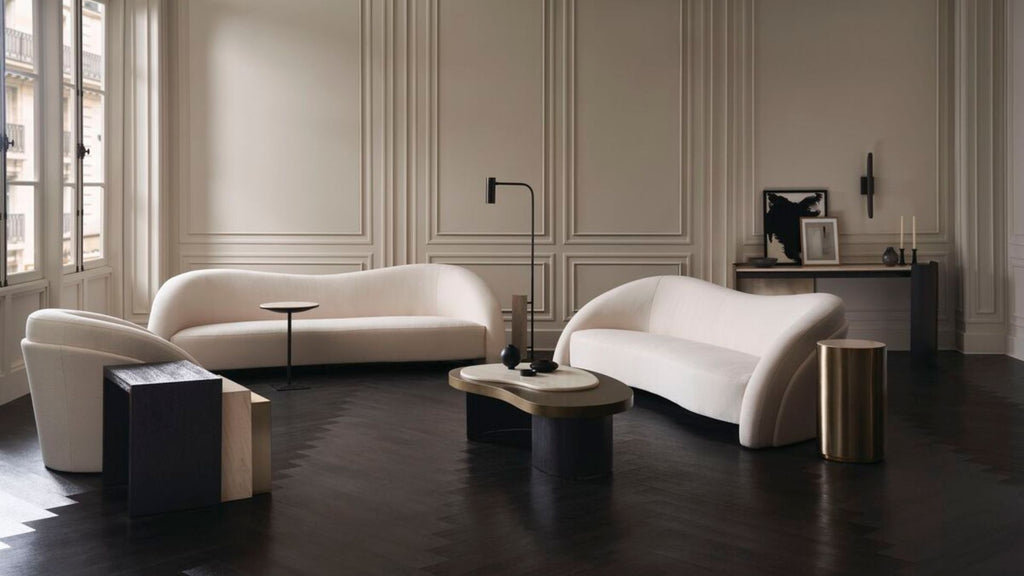 A luxury living room featuring two elegant curved white sofas, a matching armchair, a low oval coffee table, and modern side tables on a dark wood floor against a beige paneled wall.