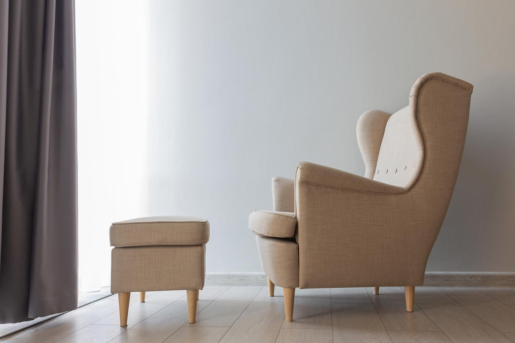 Beige linen wingback accent chair with button detail and matching ottoman on light wood legs, shown in profile against a neutral wall.