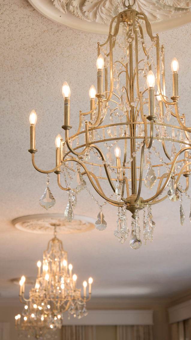 Gold-framed chandelier with curved arms and hanging crystal drops glowing with warm amber light against a pale, soft background 