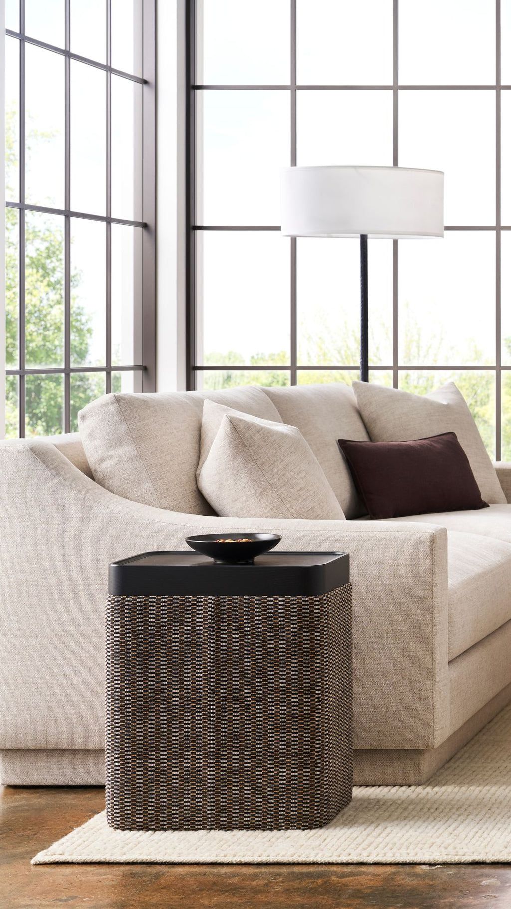 Beige sofa with a dark woven side table in a living room with large windows.