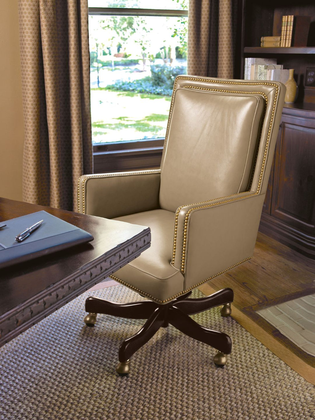 A warm taupe leather swivel office chair with gold nailhead trim and a dark wood five-star base, positioned in a home office setting next to a wooden desk.