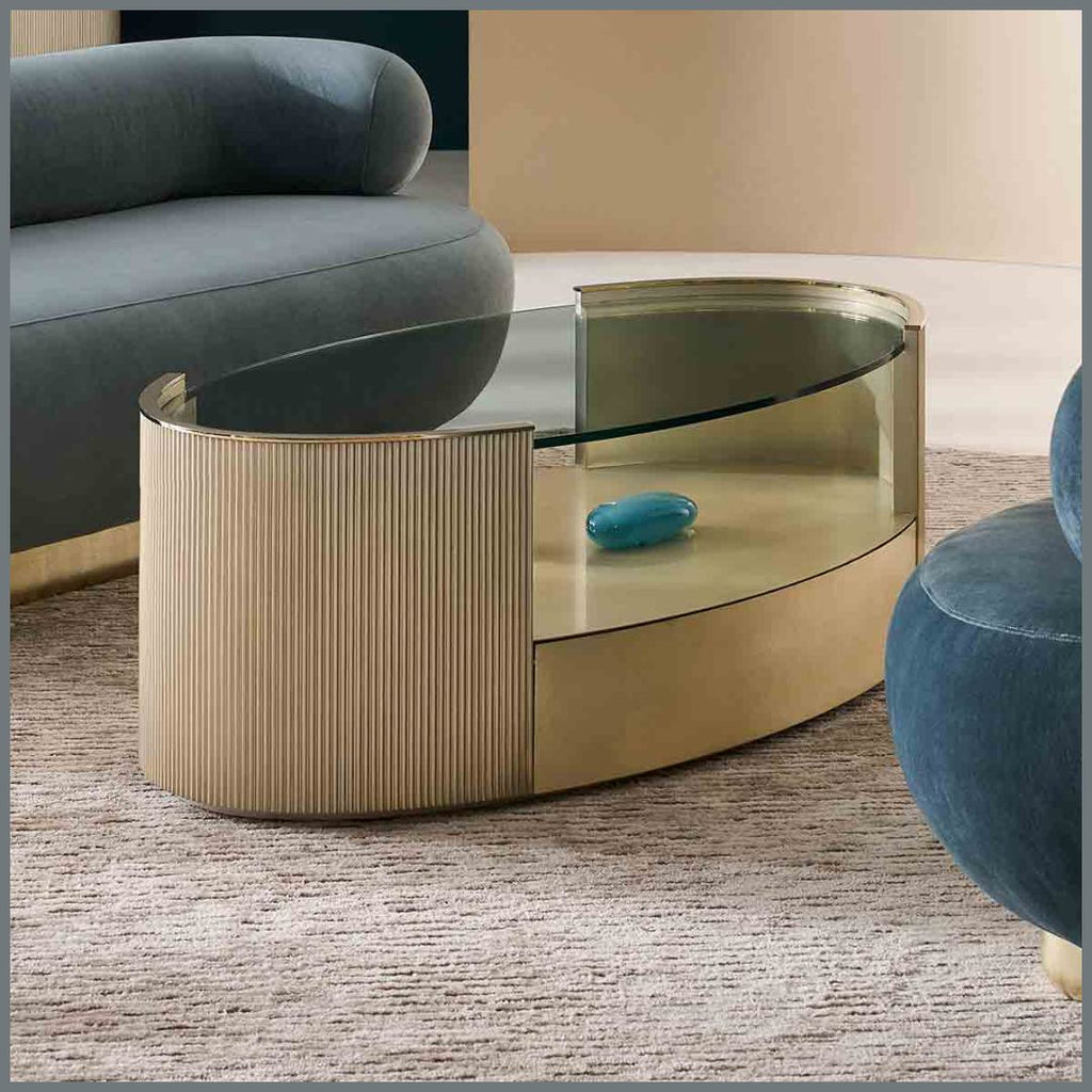 A modern curved coffee table with a ribbed metallic gold base, a clear glass top, and a glass side panel, shown in a styled living room setting.