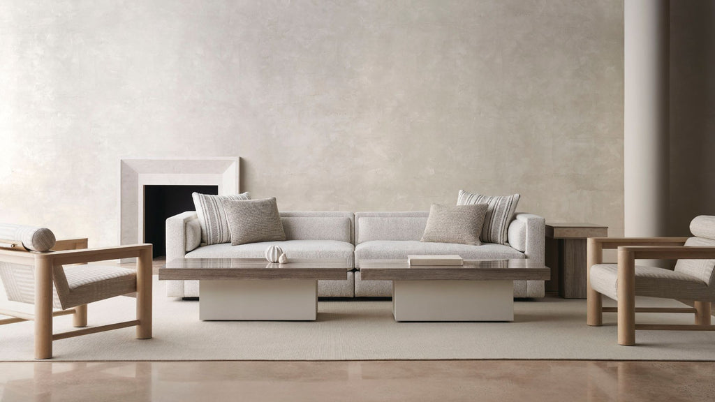A modern living room setup featuring a light gray fabric sofa with neutral throw pillows, two matching wooden armchairs, low rectangular wooden coffee tables, and a textured beige wall in the background.