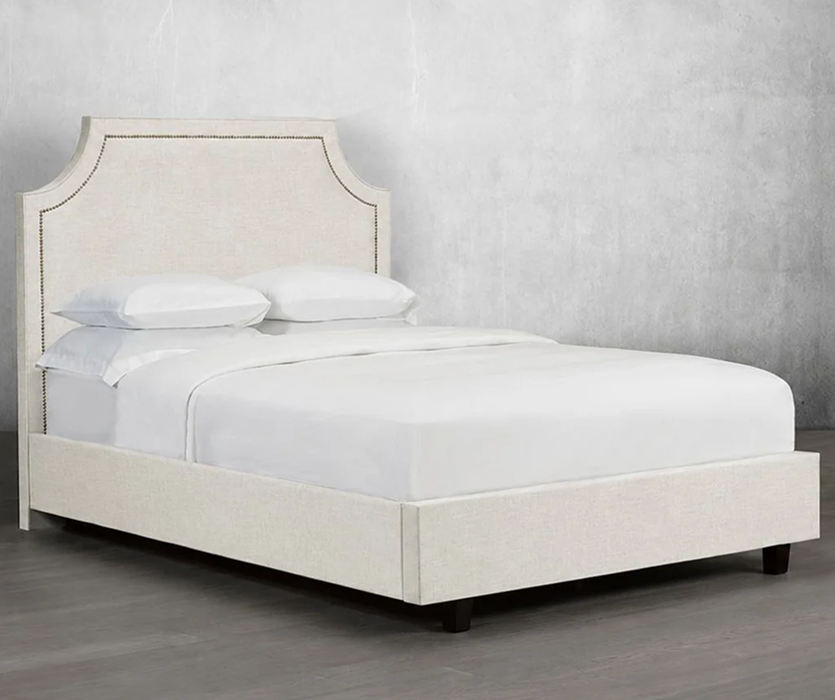 Cream upholstered bed with a tall, curved headboard edged in studded trim, shown from the front against a clean white background